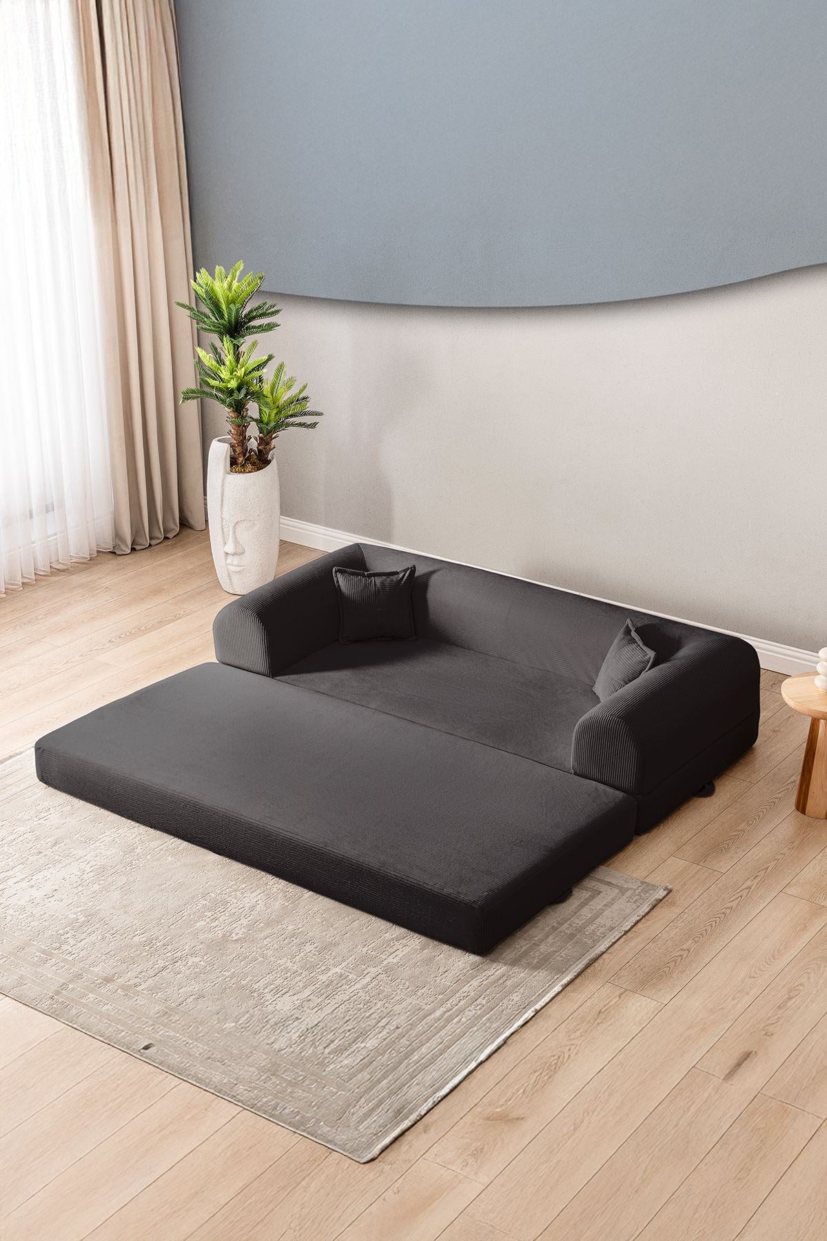 Anthrazitfarbenes 3-Sitzer Schlafsofa aus geripptem Cord-Plüschstoff mit zwei Kissen und sichtbarer rutschfester Sohle.