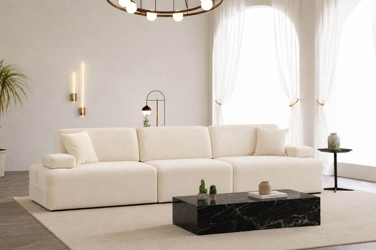 Cremefarbenes Madlen 4-Sitzer Sofa von Hanah Home mit niedrigen Kunststoffbeinen, Polyesterbezug und zwei Kissen auf hellem Untergrund.