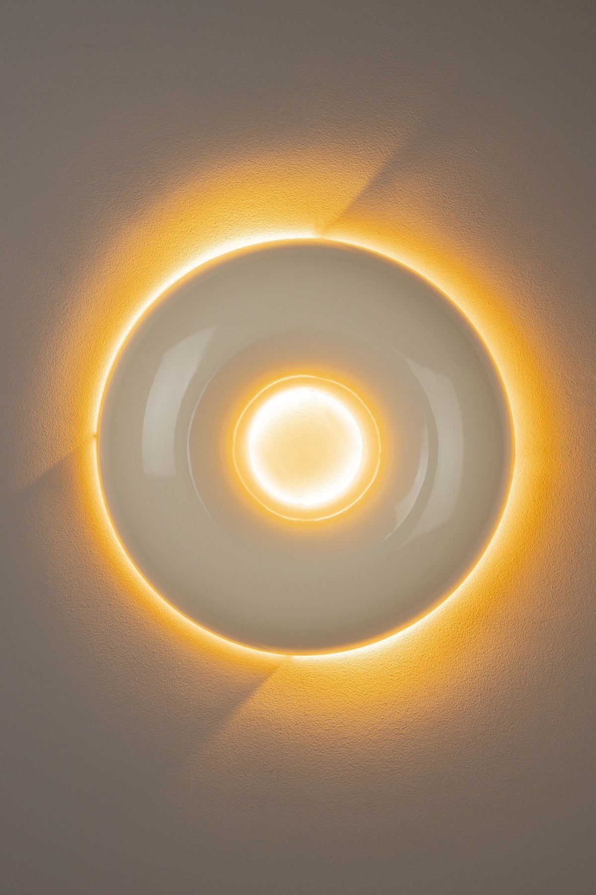 Runde beige Wandleuchte aus glänzendem Metall in Donut-Form mit indirektem Lichtaustritt entlang des Randes, montiert an einer Wand.
