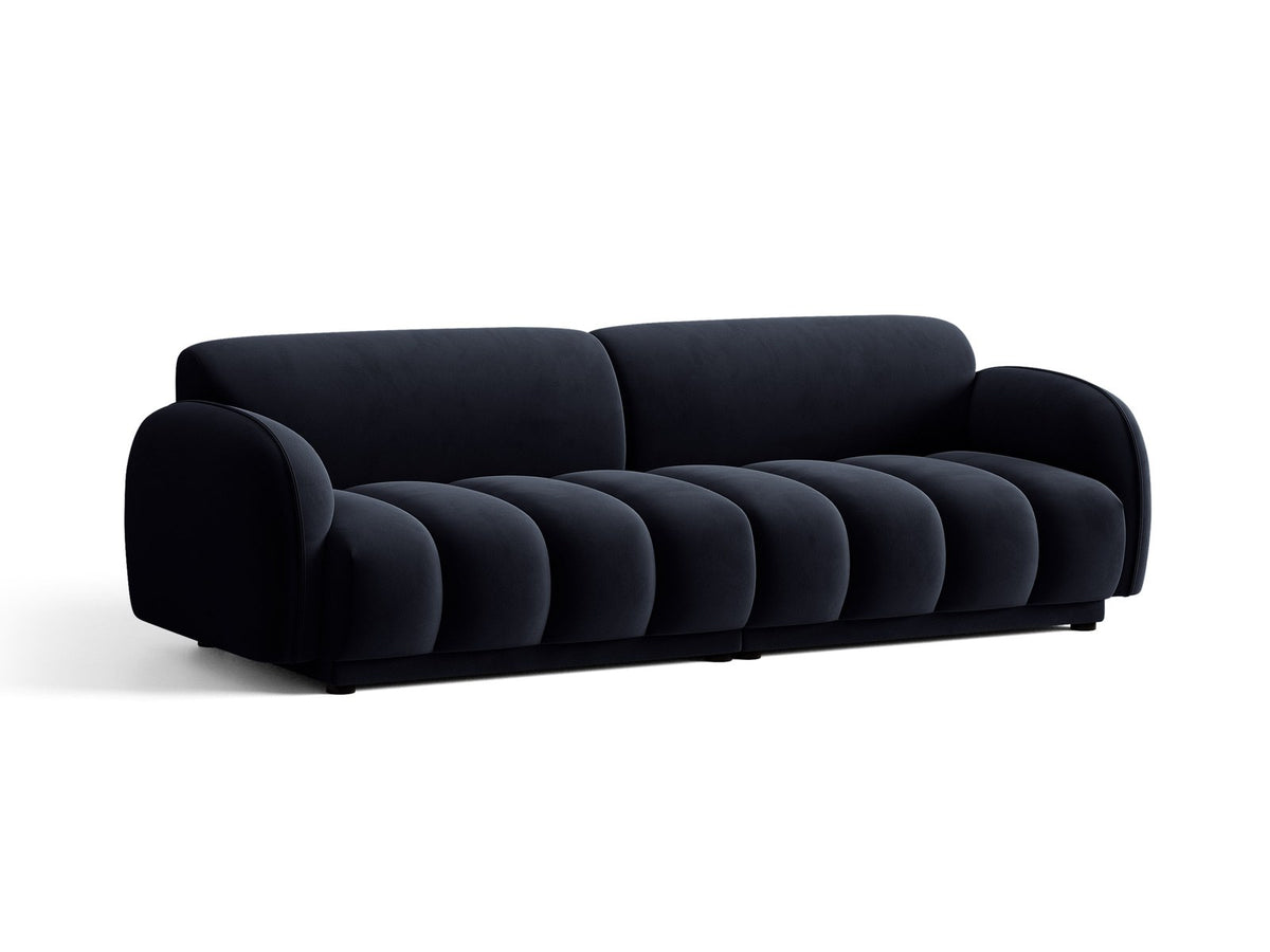 Vierersofa Bilbao mit hellbeigem Samtbezug, schwarzen Kunststoffbeinen und klarer, gerader Linienführung, fotografiert freistehend im Raum.