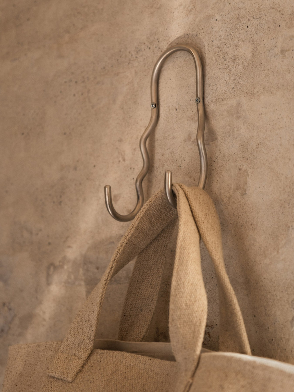 Doppelhaken Curvature von Ferm Living aus Edelstahl mit geschwungener, skulpturaler Form, montiert an einer Wand.