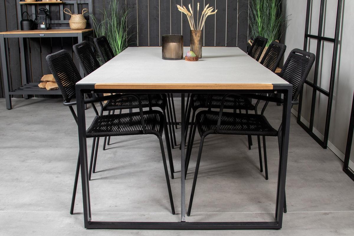 Zwei schwarze Esszimmerstühle mit geflochtener Seilsitzfläche und Aluminiumgestell neben rechteckigem Esstisch mit Glas-Teak-Platte und Stahlbeinen.