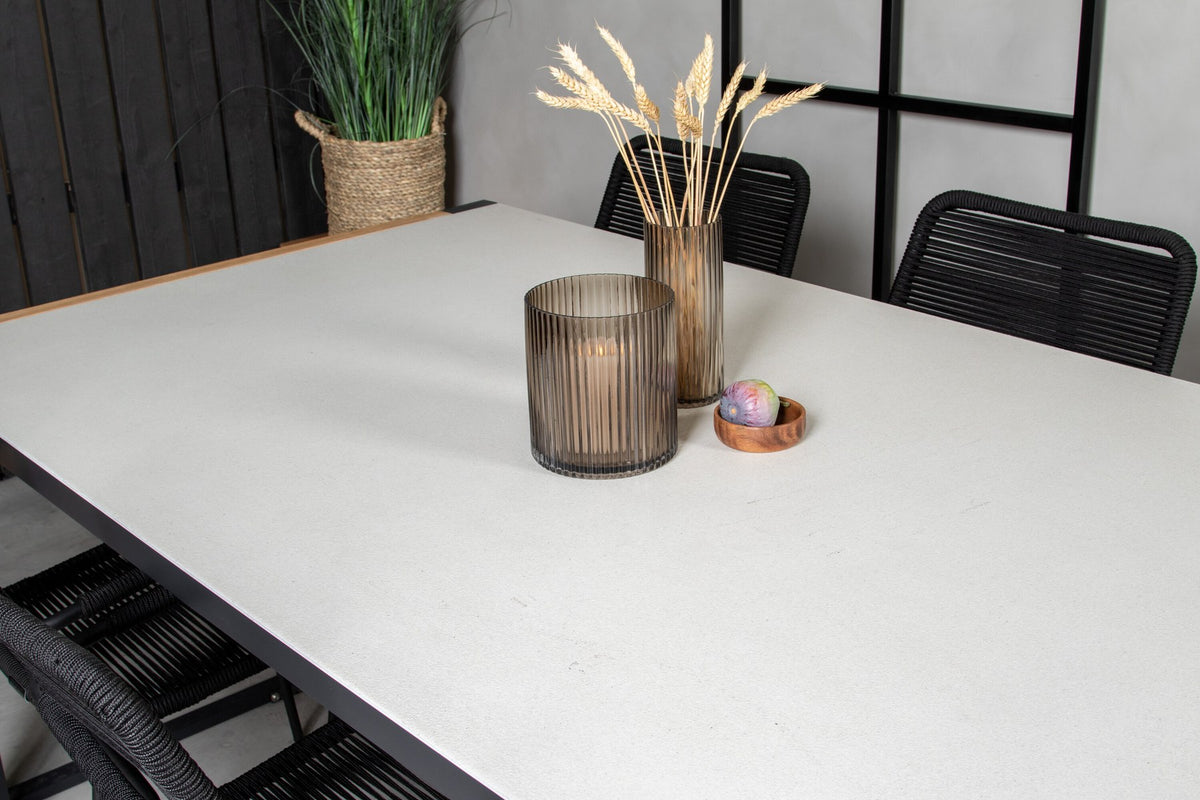 Zwei schwarze Esszimmerstühle mit geflochtener Seilsitzfläche und Aluminiumgestell neben rechteckigem Esstisch mit Glas-Teak-Platte und Stahlbeinen.