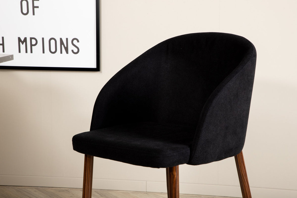 Zwei schwarze Esszimmerstühle mit Nussbaum-Beinen und Polyester-Sitzfläche neben rundem Esstisch Ø110cm mit schwarzer MDF-Platte und Stahlbeinen.