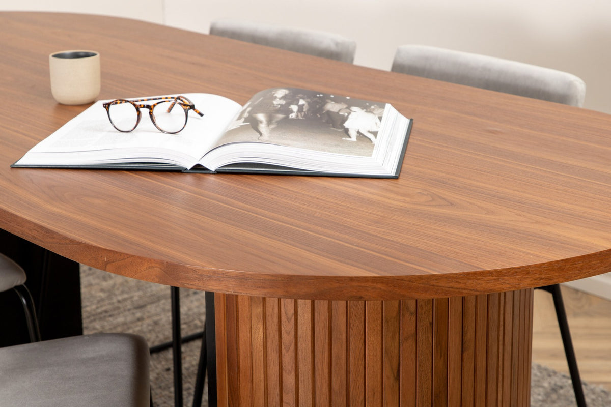 Zwei graue Esszimmerstühle mit schmalen Metallbeinen und samtigem Sitz neben ovalem Esstisch aus Nussbaum-MDF mit Furnier, 200x90cm.