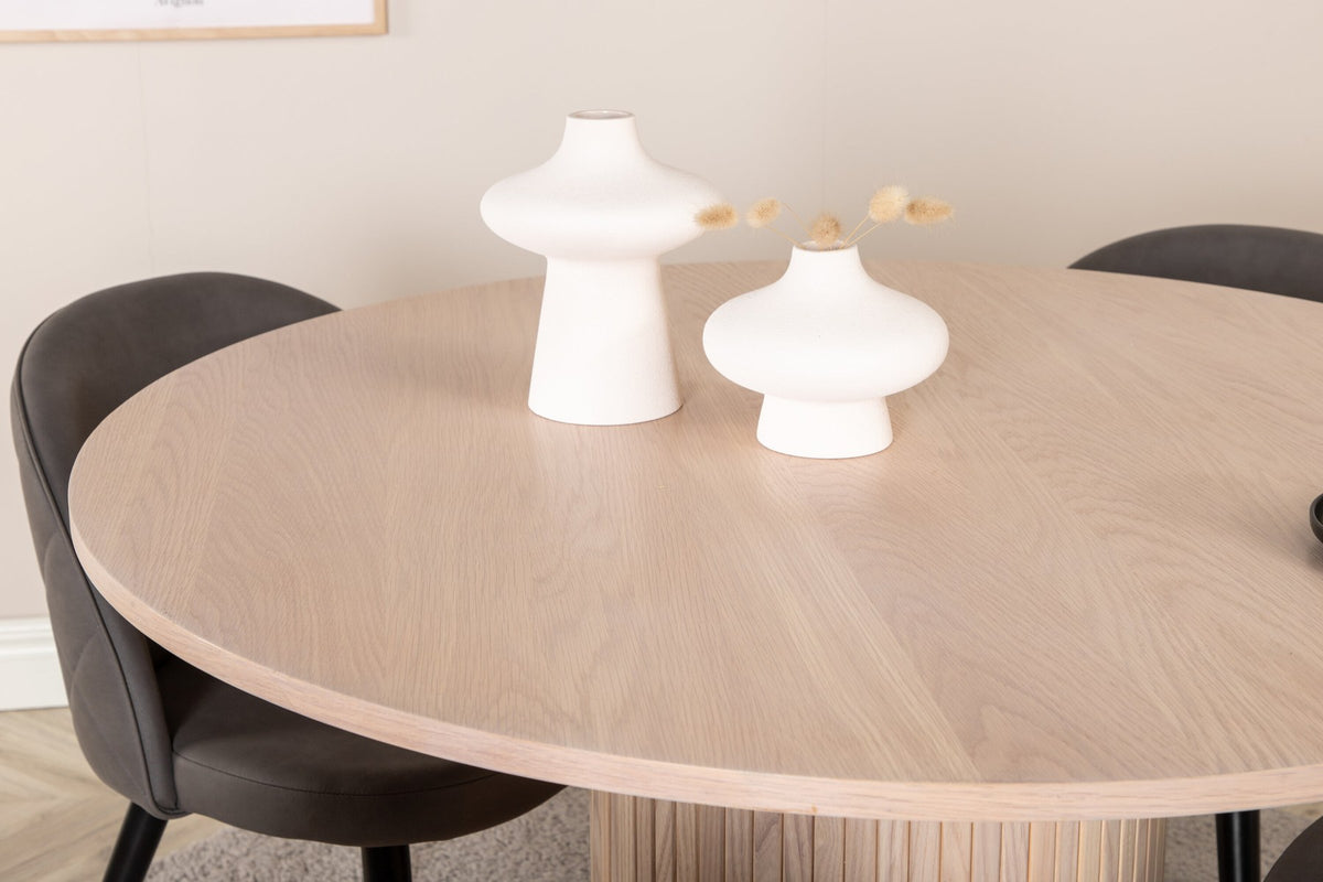 Runder Esstisch aus MDF in Whitewash mit ⌀110cm neben zwei Esszimmerstühlen mit grauem Mikrofaserstoff und schwarz pulverbeschichteten Eisenbeinen.