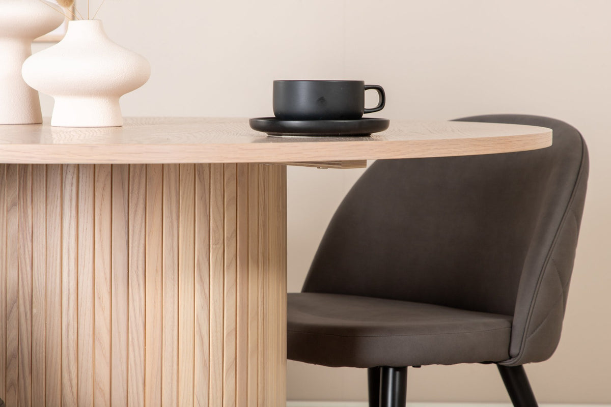 Runder Esstisch aus MDF in Whitewash mit ⌀110cm neben zwei Esszimmerstühlen mit grauem Mikrofaserstoff und schwarz pulverbeschichteten Eisenbeinen.