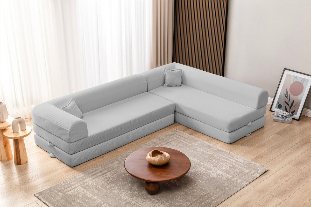 Hellgraues Ecksofa-Bett aus geripptem Cord-Plüschstoff mit niedriger Rückenlehne, vier Kissen und ausklappbarer Liegefläche.