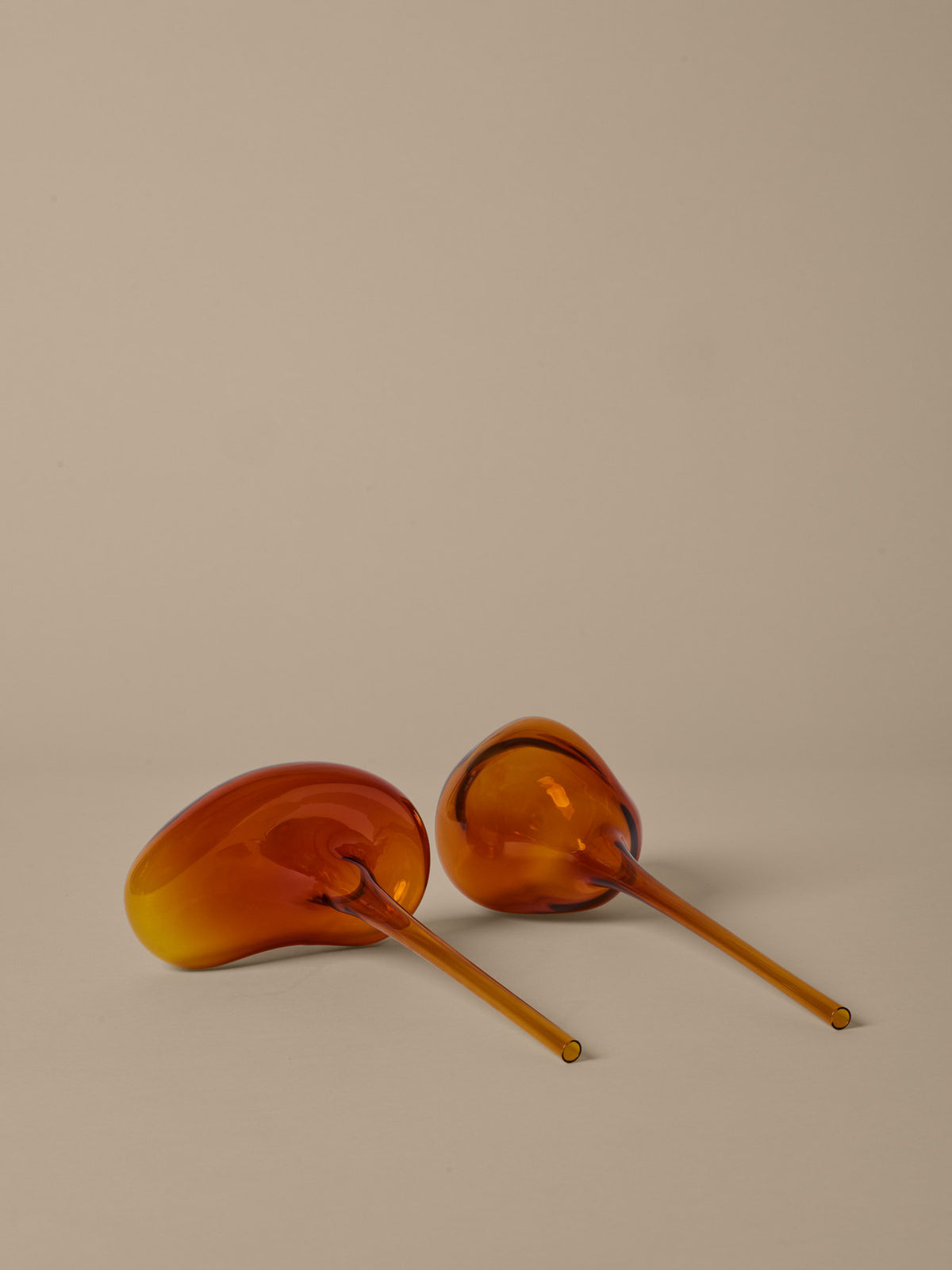 Zwei Wasserkugeln aus mundgeblasenem Glas in Amber von Ferm Living, organische Kugelform mit langem Stiel, stehend neben einem Pflanzentopf.