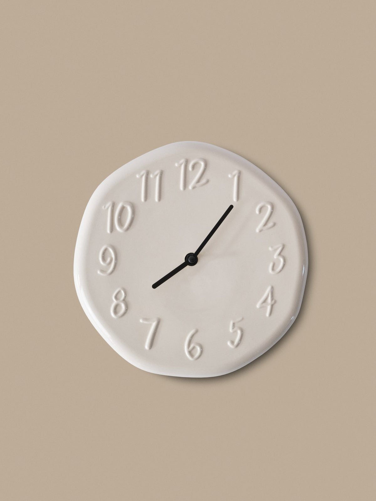 Runde Wanduhr aus glasiertem Steinzeug in Creme mit Aluminium-Zeigern und direkt in die Oberfläche modellierten Ziffern, leicht unregelmässige Form.