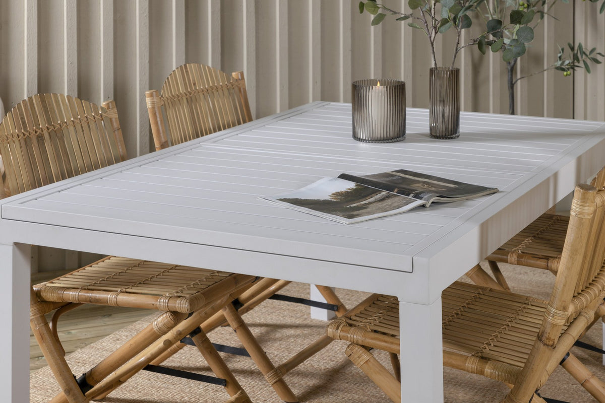 Aluminium-Caféstuhl in Natur mit Baumwoll-Polyester-Bezug neben rechteckigem Esstisch mit weisser Aluminium-Tischplatte und Rattanbeinen.