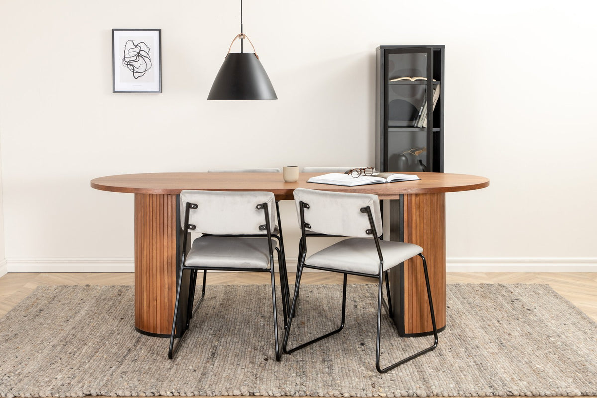 Zwei graue Esszimmerstühle mit schmalen Metallbeinen und samtigem Sitz neben ovalem Esstisch aus Nussbaum-MDF mit Furnier, 200x90cm.