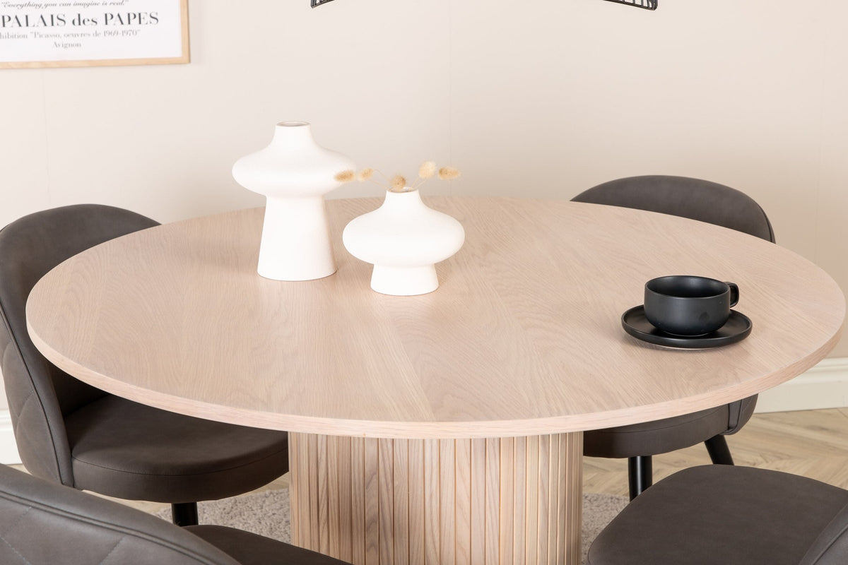 Runder Esstisch aus MDF in Whitewash mit ⌀110cm neben zwei Esszimmerstühlen mit grauem Mikrofaserstoff und schwarz pulverbeschichteten Eisenbeinen.
