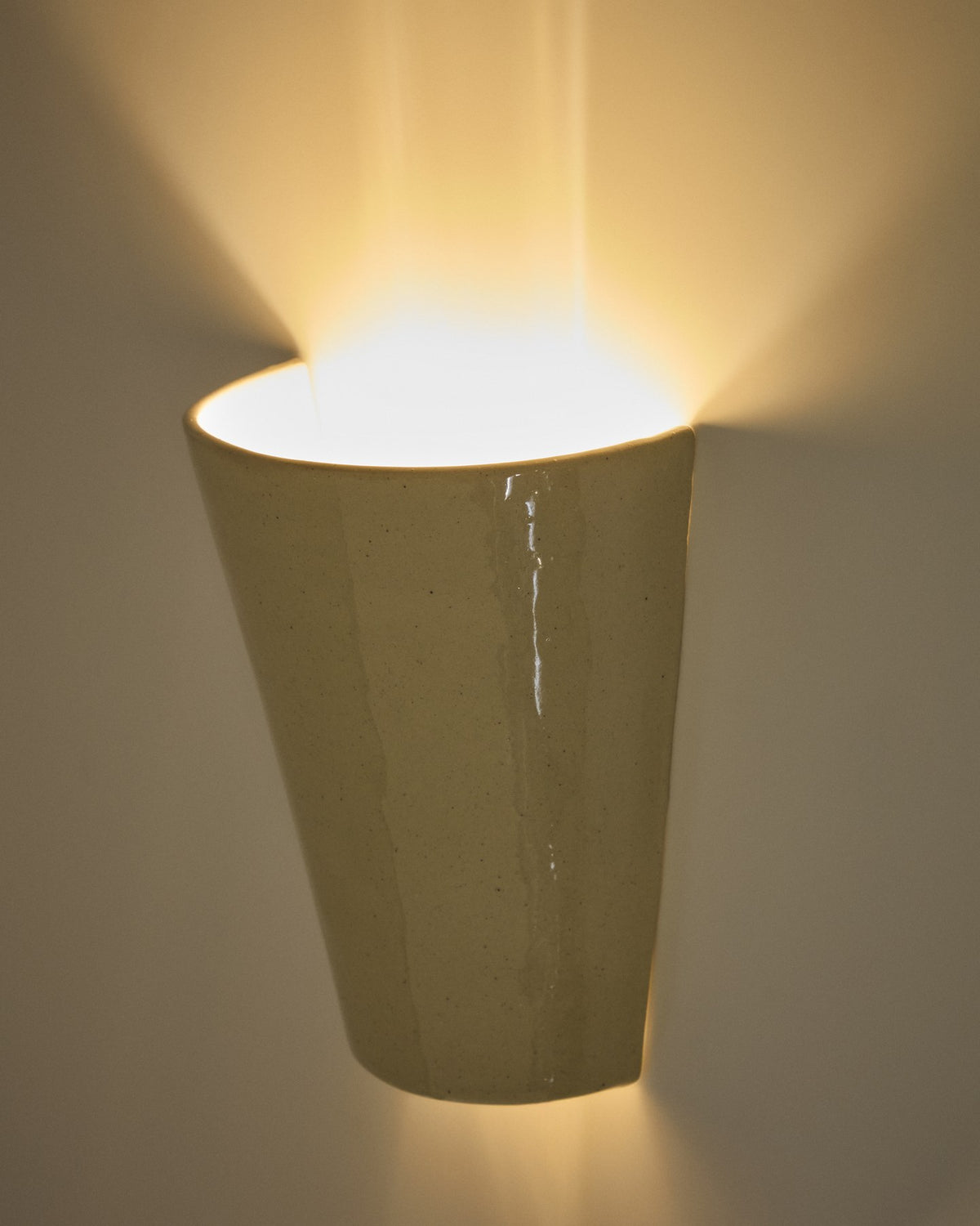 Beigefarbene Wandleuchte Ceren aus Keramik mit schlichter, runder Form, montiert an einer hellen Wand, Grösse 12 x 6 x 14 cm.