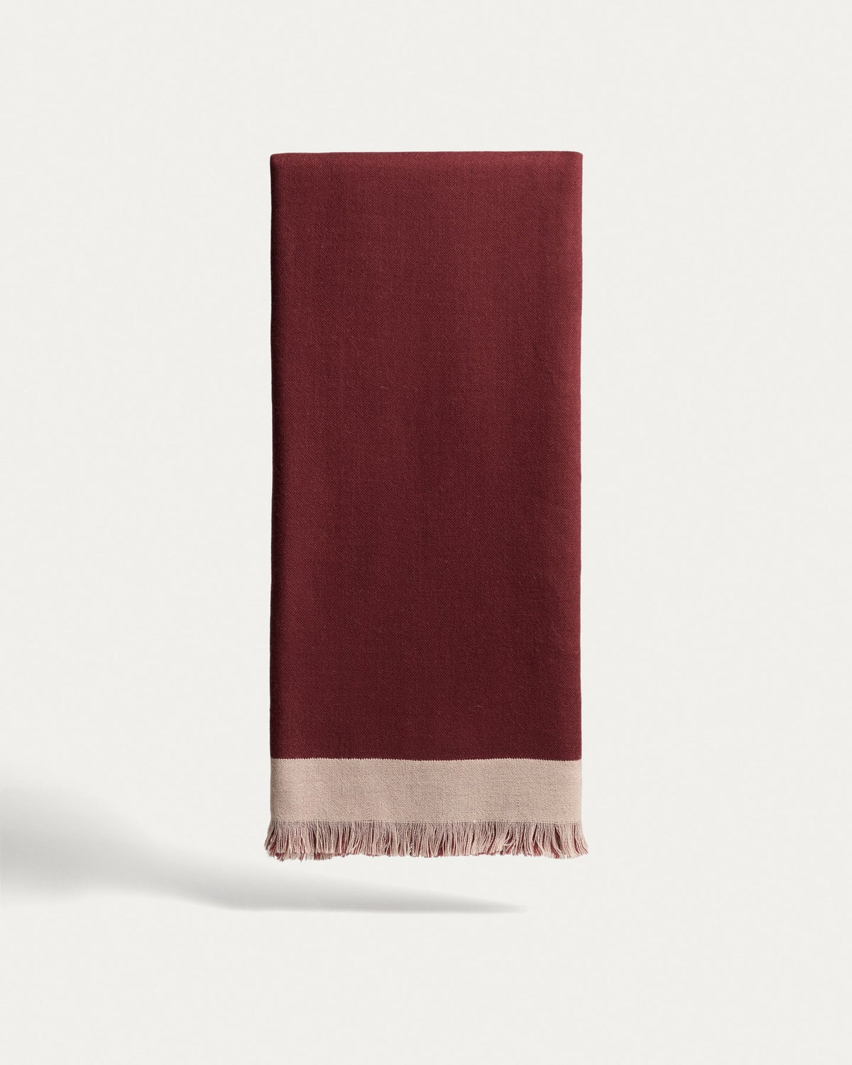 Wendbare Wolldecke aus 100% Wolle in Granatrot und Beige mit Fransen an den Rändern, gefaltet auf einem Sofa liegend.