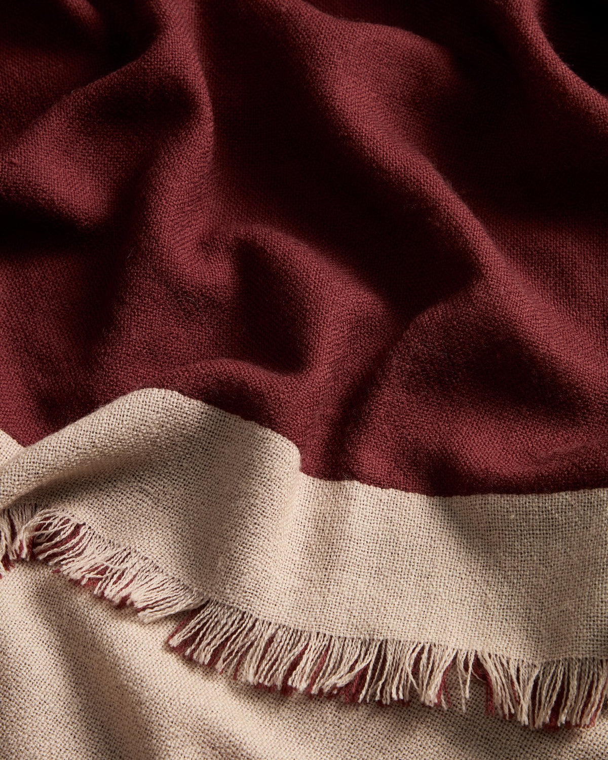Wendbare Wolldecke aus 100% Wolle in Granatrot und Beige mit Fransen an den Rändern, gefaltet auf einem Sofa liegend.